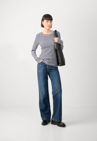 Gestreiftes langärmeliges Oberteil, navy und weiß, kombiniert mit lockeren blauen Jeans. Das Model hält eine schwarze Tote-Tasche und steht vor einem hellen Hintergrund.
