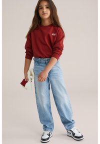 Felpa rossa con testo bianco, jeans baggy azzurri chiari e sneakers in bianco e nero. Tiene in mano un cappello lavorato a maglia bianco con dettagli rossi.