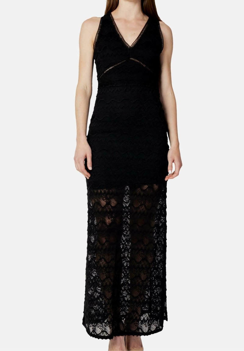 Femme portant une robe en dentelle noire sans manches avec un décolleté en V et une jupe partiellement transparente s'étendant jusqu'aux chevilles.