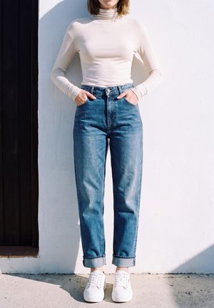 Une personne porte un jean bleu retroussé, un haut blanc à col montant et des baskets blanches. Elle se tient les mains dans les poches devant un mur ensoleillé.