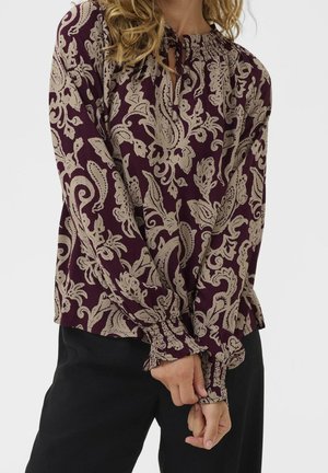 Vrouw draagt een paarse blouse met lange mouwen met beige bloemige paisley patronen en gerimpelde manchetten, gecombineerd met zwarte broek.