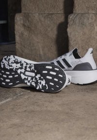 Chaussures de course Adidas blanches et noires avec semelles texturées, une chaussure debout et l'autre montrant sa semelle à motifs sur une surface en pierre.