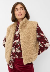 Vriendelijke beige mouwloze vest met een hoge kraag, gecombineerd met een rode en gemusterde lange mouwen shirt en bordeauxrode broek. Zachte textuur overal.