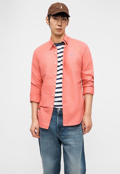 Mladić koji nosi smeđu kapu, koraljnu košulju na kopčanje preko tamnoplave i bijele prugaste majice, te široke plave traperice, stoji ispred jednostavne pozadine.