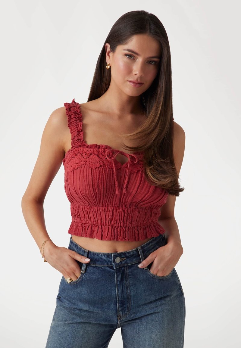 Rode cropped top met gerimpelde textuur, kantafwerking en verstelbare strikhals. G gedragen met hoog-taille blauwe jeans.