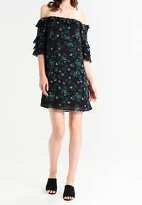 Black off-the-shoulder dress with layered ruffle sleeves, featuring a floral pattern in green and white. Worn with black open-toe mules.