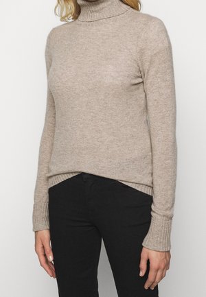Femme portant un pull à col roulé beige et un pantalon noir, debout devant un fond blanc uni.