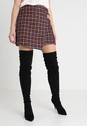 Femme portant une mini-jupe en tweed multicolore avec un ourlet asymétrique et des bottes cuissardes en daim noir à talons hauts.