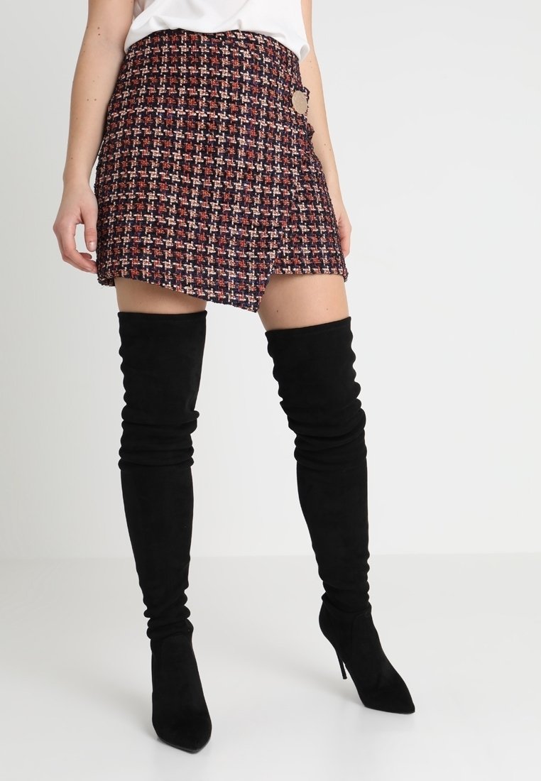 Femme portant une mini-jupe en tweed multicolore avec un ourlet asymétrique et des bottes cuissardes en daim noir à talons hauts.