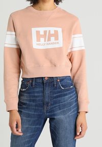 Felpa cropped rosa con strisce bianche, caratterizzata da un logo quadrato bianco "HH" e "HELly HANSEN". Indossata con jeans in denim a vita alta.
