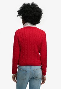 Rode kabelgebreide trui met geribbelde zoom en manchetten, gedragen over een wit overhemd met kraag, gecombineerd met lichtblauwe spijkerbroek. Achteraanzicht weergegeven.