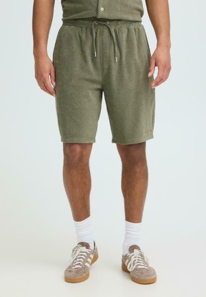 Hombre con pantalones cortos de felpa verde oliva con cordón, calcetines blancos y zapatillas de ante beige, de pie contra un fondo blanco.