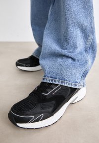 Zapatillas deportivas negras con paneles de malla y suelas de goma blancas, combinadas con jeans de mezclilla azul claro, mostrando un ajuste relajado.