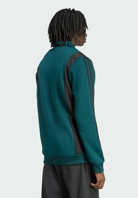 Teal zip-jakke med sorte sidepaneler og tre sorte striber på ærmerne; stoffet ser glat og åndbart ud med en afslappet pasform.