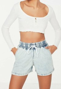 Valkoinen ribattu crop-top, jossa on avara kaula-aukko ja kolme nappia, yhdistettynä vaaleansinisiin farkkushortseihin, joissa on joustava vyötärö ja naru.
