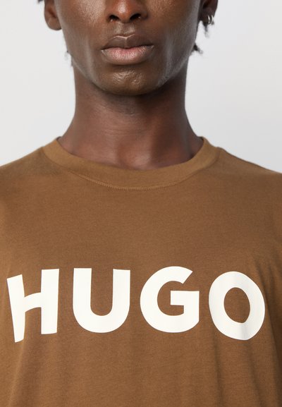Крупен план на човек, носещ кафява риза с големи бели букви, изписващи "HUGO" на гърдите.