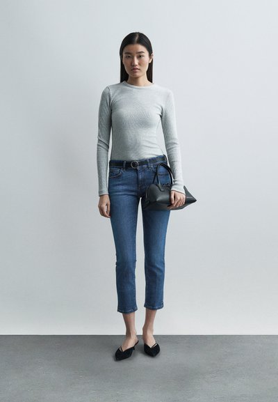 Une femme se tient contre un mur uni, portant un haut gris à manches longues, un jean bleu raccourci, des chaussures pointues noires, tenant un sac à main noir dans la main droite.