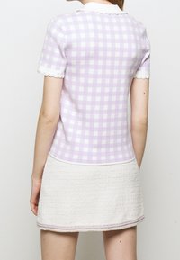 Kobieta w krótkim rękawie lawendowej i białej koszulce w kratkę z białym kołnierzykiem oraz teksturowanej kremowej spódnicy mini z lawendowym wykończeniem.