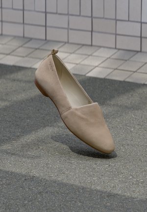 Beige suède platte schoen met puntige neus zwevend boven geplaveide grond tegen een betegelde muur als achtergrond.
