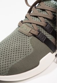 Grön textil-sneaker med ovandel i mesh, bruna snörningar, svarta detaljer och en vit sula. Har "EQT"-logotypen på tungan.