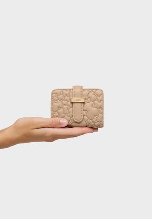 Ręka trzymająca mały beżowy portfel z tłoczonymi wzorami kwiatów i paskiem z zapięciem w odcieniu złota na jednolitym jasnym tle.