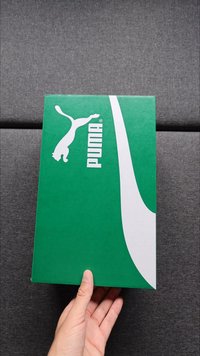 Roheline pappkarp, millel on valge Puma logo ja swoosh-disain. Pinna kohal on sile ja matt viimistlus. Käsi hoiab karpi.