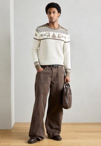 Krémový pletený sveter so vzormi hnedých sobov a vločiek; v kombinácii s voľnými hnedými nohavicami; drží hnedú tašku; topánky sú tmavohnedé.