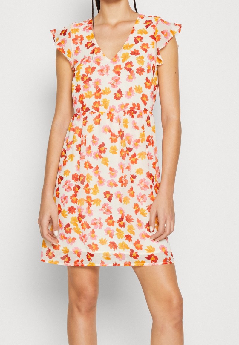 Femme portant une robe blanche sans manches avec des motifs floraux rouges, orange et roses et des manches volantées, debout devant un fond uni.