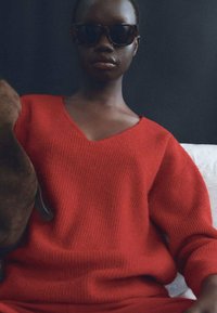 Maglione a coste rosso con scollo a V. Modello che indossa occhiali da sole scuri, seduto contro uno sfondo scuro. Dettaglio del tessuto testurizzato visibile.