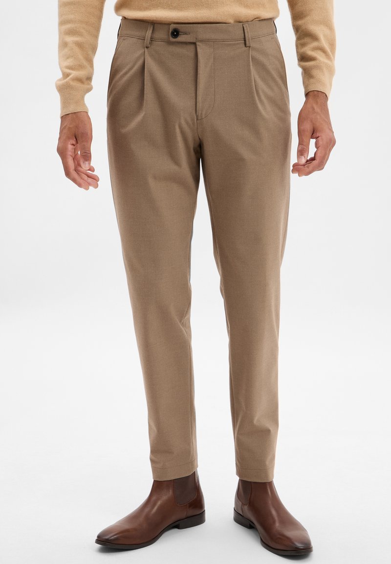 Beige tailored trousers with pleats, featuring a button closure and belt loops. Worn with brown leather ankle boots. Smooth texture.