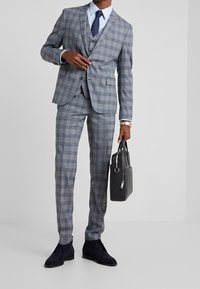 Uomo con indosso un completo a quadri grigi e una cravatta blu navy, che tiene una valigetta in pelle nera nella mano destra, su uno sfondo chiaro e uniforme.