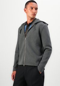 Grå zip-up hettegenser med hette, lange ermer og en myk tekstur. Har en frontglidelås og bæres over en blå og brun polo skjorte.