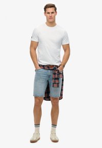 Hvid kortærmet t-shirt parret med blå denimshorts, en rød og sort rutet skjorte bundet om taljen, og beige sneakers.
