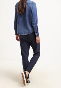 Chemise en denim bleue à l'ourlet arrondi et à poignets à boutons, associée à un jogger marine avec des accents sur les côtés et des baskets noires.