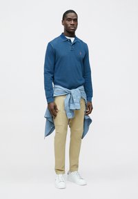 Polo azul de manga comprida com colarinho, camisa azul-claro presa na cintura, calças cáqui e ténis brancos. Fundo simples.