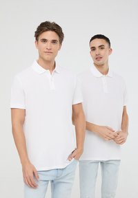 Due uomini indossano magliette polo bianche a maniche corte con colli bottonati, abbinate a jeans in denim azzurro chiaro. Il tessuto appare liscio e uniforme.