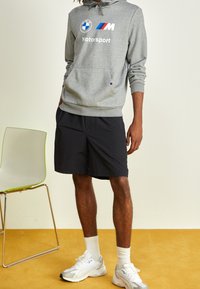 Sudadera gris con capucha y el logo "BMW Motorsport", combinada con pantalones cortos negros, calcetines blancos y zapatos atléticos blancos sobre una alfombra amarilla.