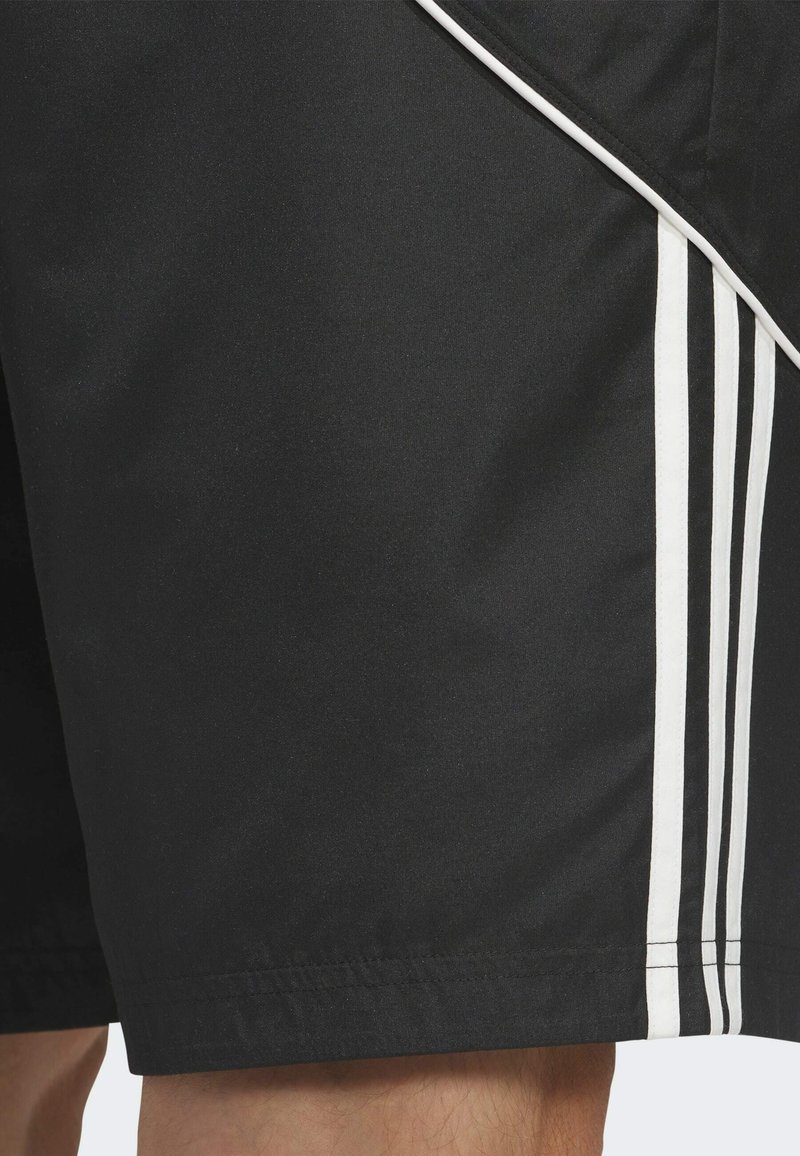 Short noir avec trois bandes blanches verticales sur le côté, porté sur la jambe d’une personne, montrant la texture du tissu et les détails de couture.
