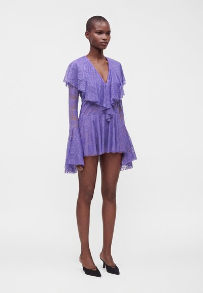 Femme portant une robe courte en dentelle violette avec des manches longues évasées, un décolleté en V profond et des volants superposés, associée à des talons noirs pointus.