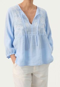 Blusa di lino azzurra chiara con maniche a tre quarti, scollatura a V e ricamo floreale bianco sul petto, caratterizzata da una vestibilità rilassata.