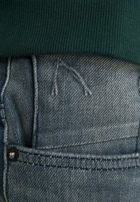 Džínové kalhoty mají tmavě modrou barvu, viditelné švy a zadní kapsu. Vrchol kalhot je částečně překrytý zeleným svetrem.