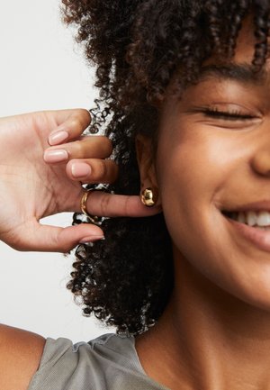 Mujer sonriente con cabello rizado toca su pendiente dorado con una mano, lleva un anillo de oro y una camiseta sin mangas gris.