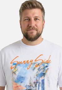 Hombre con cabello corto rubio y barba que lleva una camiseta blanca con una ilustración colorida de un paisaje tropical costero y el texto naranja "Sunny Side".