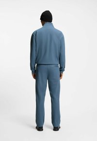 Hellblauer Trainingsanzug mit vertikaler Rippung, bestehend aus einer Jacke mit Reißverschluss am Kragen und einer Hose mit elastischem Bund, kombiniert mit einer schwarzen Mütze.