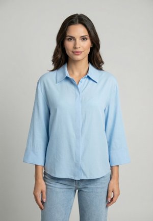 Femme aux cheveux foncés portant une chemise bleu clair boutonnée à manches trois-quarts et un jean bleu clair, debout devant un fond gris uni.