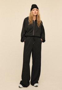 Mustaraidallinen verkkatakki, jossa on vetoketjulla suljettava huppari ja väljäleikkaiset housut, valmistettu pehmeästä materiaalista. Koristeltu sivutaskuilla.