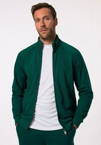 Giacca verde con zip e colletto alto, caratterizzata da una texture di tessuto liscia e un logo discreto sul petto, abbinata a una maglietta bianca a girocollo.