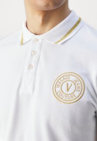 Homme portant un polo blanc avec des bordures dorées au col et un logo Versace Jeans Couture brodé en or sur la poitrine.