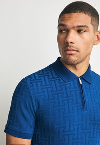 Blauw polohemd met korte mouwen met een gestructureerd geometrisch patroon, voorzien van een ritssluiting en een effen kraag. Gladde stofafwerking.