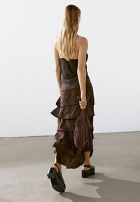 Gonna a strati marrone con bordo arricciato, abbinata a un top nero senza maniche. Tiene una borsa marrone con texture e indossa scarpe nere chunky.